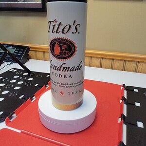 May include: A white travel mug with a black lid and a gold bottom. The mug has a Tito's Handmade Vodka logo with a red star and the words "Austin, Texas".