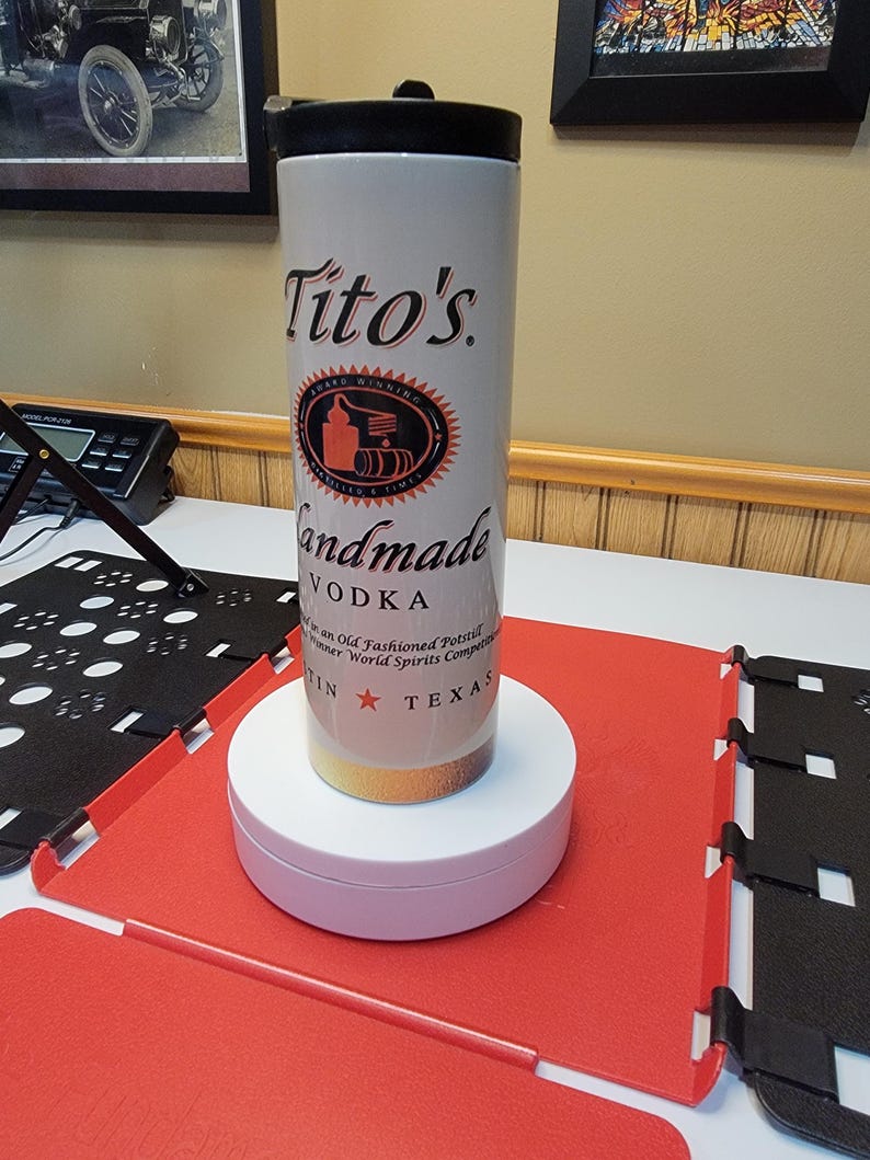 May include: A white travel mug with a black lid and a gold bottom. The mug has a Tito's Handmade Vodka logo with a red star and the words "Austin, Texas".