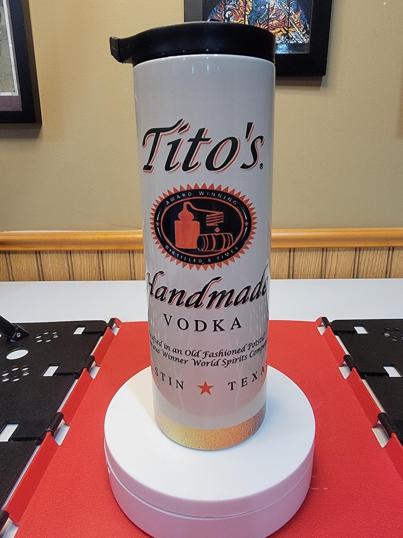 May include: A white travel mug with a black lid and a gold band around the bottom. The mug has a logo for Tito's Handmade Vodka, which includes the text "Tito's", "Award Winning", "Distilled 6 Times", "Handmade Vodka", and "Austin, Texas".
