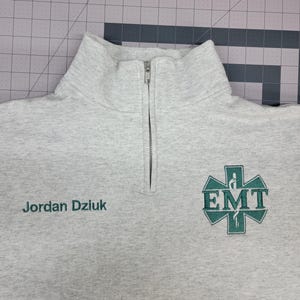 May include: A light grey quarter-zip sweatshirt with a high collar. The sweatshirt features the embroidered text "Jordan Dziuk" in green on the left chest and a green EMT logo on the right chest.