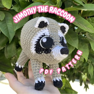 May include: A crocheted raccoon toy featuring a light brown body, white accents on the face and ears, and black patches around the eyes, nose, and paws. The text JIMOTHY THE RACCOON and PDF PATTERN is overlaid in pink on the image.