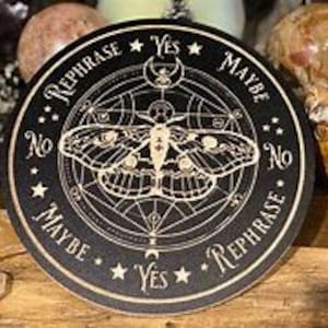 Può includere: Un vassoio rotondo in legno nero con un design geometrico dorato e un'illustrazione di una falena. Il vassoio ha le parole "Sì", "Forse", "No" e "Riformulare" scritte intorno al bordo.