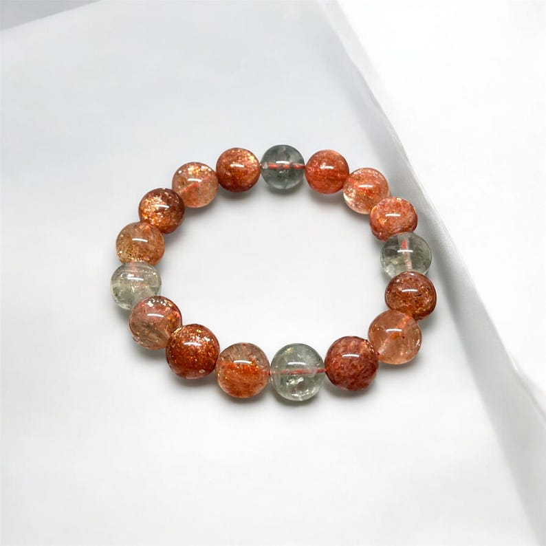 K&ouml;nnte beinhalten: Ein Armband mit polierten, runden Edelsteinen in transparenten Gr&uuml;nt&ouml;nen und r&ouml;tlich-braunen Farbt&ouml;nen. Die Perlen weisen funkelnde Einschl&uuml;sse auf. Das Armband ist auf einem elastischen Band aufgef&auml;delt und wirkt nat&uuml;rlich und erdig.