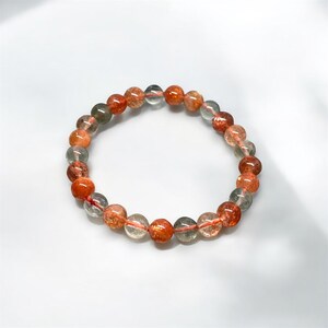 K&ouml;nnte beinhalten: Ein Armband aus runden, durchscheinenden Perlen in Orange-, Rot- und Klart&ouml;nen. Die Perlen sind auf einem roten Gummiband aufgef&auml;delt. Das Armband wird auf einer wei&szlig;en Oberfl&auml;che pr&auml;sentiert. Jede Perle misst etwa 1 cm im Durchmesser.
