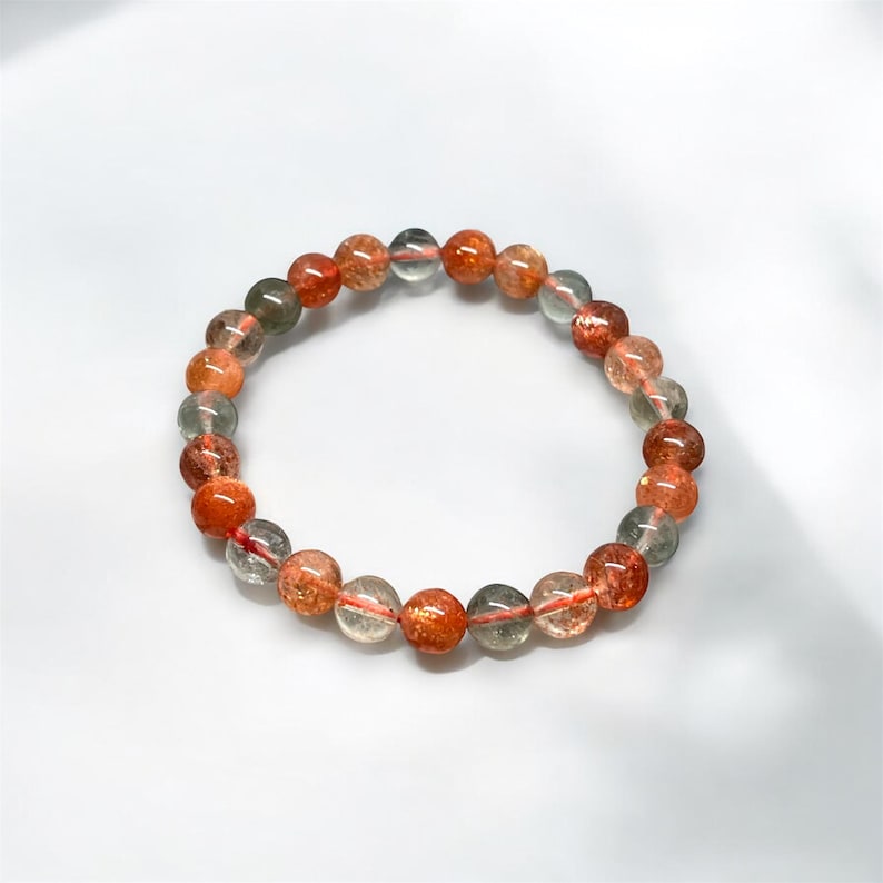 K&ouml;nnte beinhalten: Ein Armband aus runden, durchscheinenden Perlen in Orange-, Rot- und Klart&ouml;nen. Die Perlen sind auf einem roten Gummiband aufgef&auml;delt. Das Armband wird auf einer wei&szlig;en Oberfl&auml;che pr&auml;sentiert. Jede Perle misst etwa 1 cm im Durchmesser.