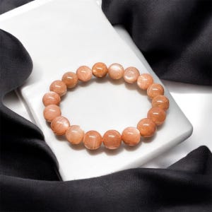 Puede incluir: Un brazalete hecho de cuentas redondas de piedra solar de color melocot&oacute;n. Las cuentas brillantes est&aacute;n ensartadas para formar un c&iacute;rculo. El brazalete se exhibe sobre una superficie blanca con un fondo de tela negra.