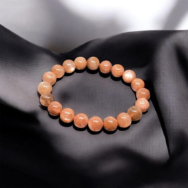 Puede incluir: Un brazalete hecho de cuentas redondas de piedra solar de color melocot&oacute;n. Las cuentas tienen un brillo sutil y est&aacute;n ensartadas para formar una forma circular. El brazalete se exhibe sobre una tela oscura y texturizada, creando un contraste.