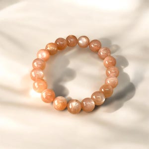 Puede incluir: Una pulsera hecha de cuentas redondas de piedras preciosas de color melocot&oacute;n. Las cuentas tienen una superficie pulida y reflectante y var&iacute;an ligeramente en color, con algunas mostrando un brillo dorado. La pulsera se muestra sobre una superficie blanca.