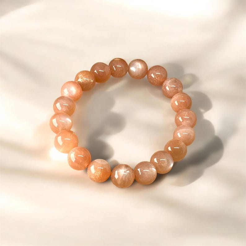 Puede incluir: Una pulsera hecha de cuentas redondas de piedras preciosas de color melocot&oacute;n. Las cuentas tienen una superficie pulida y reflectante y var&iacute;an ligeramente en color, con algunas mostrando un brillo dorado. La pulsera se muestra sobre una superficie blanca.