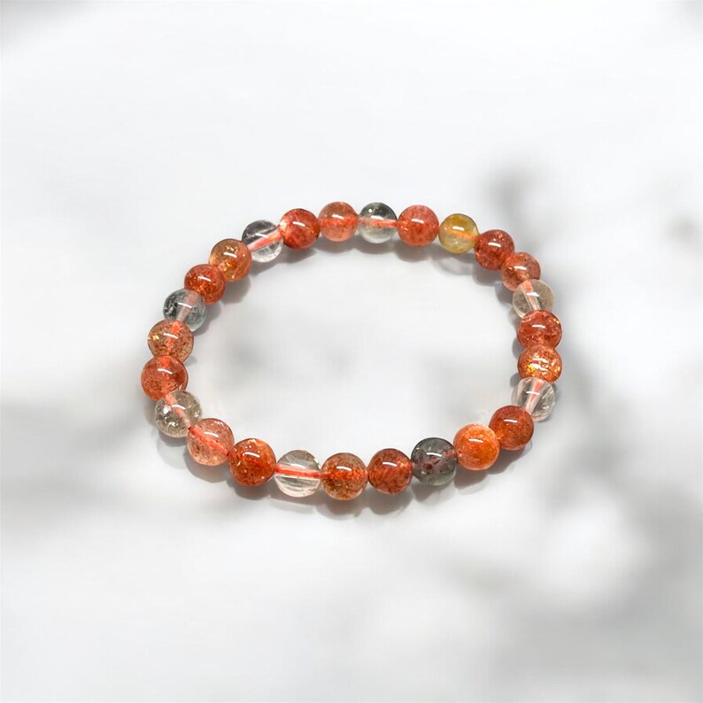 K&ouml;nnte beinhalten: Ein Armband aus orangefarbenen und durchscheinenden Perlen mit schimmerndem Effekt. Die Perlen sind auf einem d&uuml;nnen, transparenten Gummiband aufgef&auml;delt. Die Perlen variieren in Gr&ouml;&szlig;e und Farbe, einige erscheinen klar, andere orange und braun.