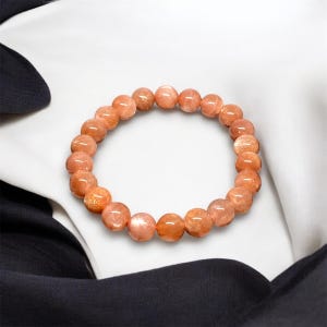 Puede incluir: Un brazalete hecho de piedras preciosas redondas y pulidas de color naranja. Las cuentas est&aacute;n ensartadas para formar una forma circular. El brazalete se muestra sobre un fondo de tela blanca y negra.