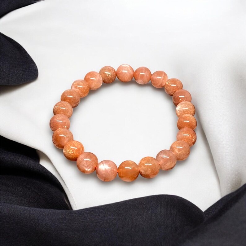 Puede incluir: Un brazalete hecho de piedras preciosas redondas y pulidas de color naranja. Las cuentas est&aacute;n ensartadas para formar una forma circular. El brazalete se muestra sobre un fondo de tela blanca y negra.