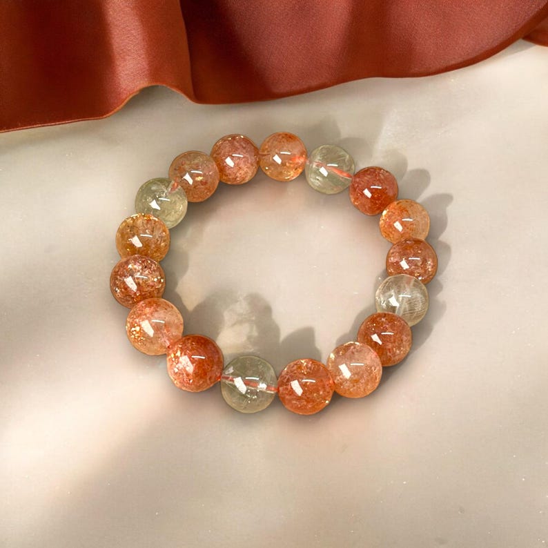 K&ouml;nnte beinhalten: Ein Armband aus runden, durchscheinenden Perlen in Orange-, Rot- und Klart&ouml;nen. Die Perlen haben ein gesprenkeltes Aussehen und sind zu einem Kreis aufgef&auml;delt. Das Armband wird auf einer hellen Oberfl&auml;che pr&auml;sentiert. Jede Perle misst etwa 1 cm im Durchmesser.