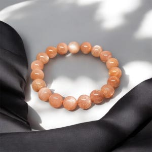 Puede incluir: Un brazalete hecho de cuentas redondas de piedra solar de color melocot&oacute;n. Las cuentas tienen un brillo sutil y est&aacute;n ensartadas para formar una forma circular. El brazalete se exhibe sobre una superficie blanca con una tela negra en el fondo.