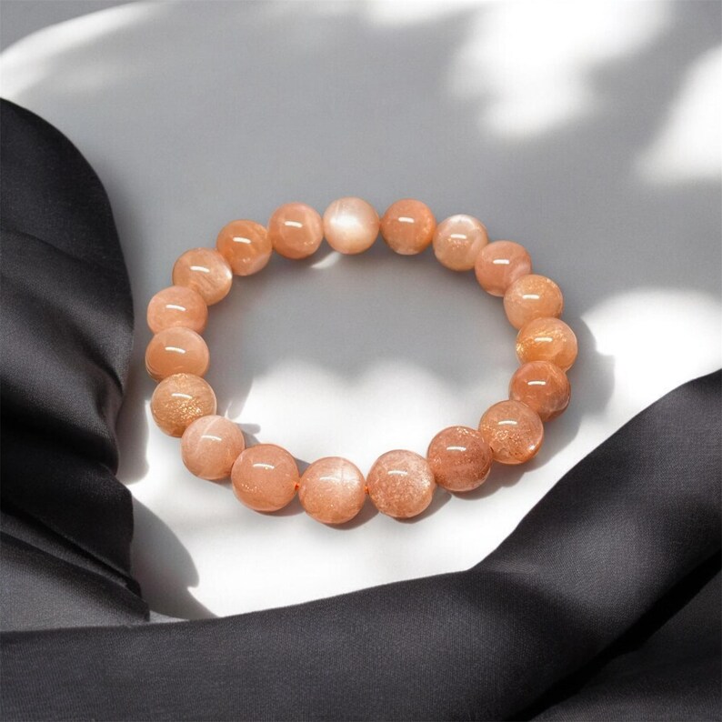Puede incluir: Un brazalete hecho de cuentas redondas de piedra solar de color melocot&oacute;n. Las cuentas tienen un brillo sutil y est&aacute;n ensartadas para formar una forma circular. El brazalete se exhibe sobre una superficie blanca con una tela negra en el fondo.