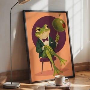 May include: A framed wall art print featuring a green frog dressed in a dark green tuxedo with a purple bow tie, sitting on a small stool. The frog holds a large green olive on a toothpick against a purple circular background and orange backdrop.