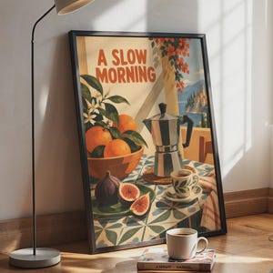 Könnte beinhalten: Ein gerahmter Kunstdruck mit dem Titel A SLOW MORNING zeigt eine mediterrane Szene mit einer Schale Orangen, einer Espressokanne, einer Kaffeetasse und aufgeschnittenen Feigen auf einer gemusterten Tischdecke vor einer Küstenkulisse.