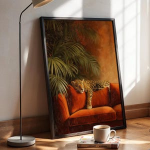 Könnte beinhalten: Ein gerahmter, rechteckiger Wandkunstdruck, der einen Leoparden zeigt, der auf einem leuchtend orangefarbenen Samtsofa unter großen grünen Palmenblättern ruht. Das Kunstwerk befindet sich in einem dünnen schwarzen Rahmen mit warmen Farbtönen.