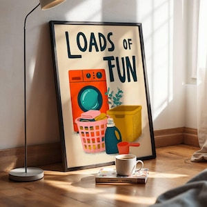 Könnte beinhalten: Ein gerahmter Kunstdruck mit dem Text "LOADS OF FUN" in dunkelblauen Buchstaben. Die Illustration zeigt eine orangefarbene Waschmaschine, einen rosa Wäschekorb mit Kleidung, einen gelben Behälter, eine blaue Waschmittelflasche und eine rote Schaufel.
