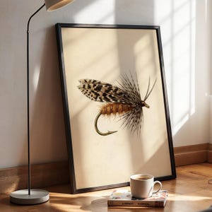 May include: A framed vertical wall art print featuring a detailed illustration of a fishing fly with brown, tan, and black speckled feathers and a gold-coloured hook, set against a plain, light cream-coloured background within a thin black frame.