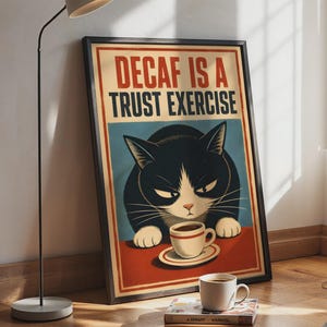 Könnte beinhalten: Ein gerahmtes, rechteckiges Wandposter mit einer schwarz-weißen Katze mit mürrischem Gesichtsausdruck, die an einem Tisch mit einer Kaffeetasse sitzt. Der Text DECAF IS A TRUST EXERCISE steht in fetten roten und schwarzen Buchstaben darüber.
