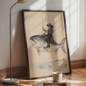 Pode incluir: Uma impressão de arte emoldurada com uma ilustração surreal de uma pessoa com chapéu de cowboy e jaqueta azul montada em um peixe grande. O cavaleiro segura um laço acima da cabeça sentado em uma sela de couro marrom sobre o peixe.