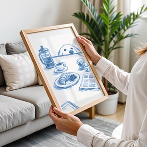May include: A rectangular wooden frame containing a white print with blue ink illustrations of a breakfast scene. The artwork depicts a French press, a coffee cup, a croissant, a soft-boiled egg, a jar, and a newspaper, with a dog peeking over the table.