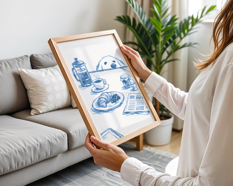 May include: A rectangular wooden frame containing a white print with blue ink illustrations of a breakfast scene. The artwork depicts a French press, a coffee cup, a croissant, a soft-boiled egg, a jar, and a newspaper, with a dog peeking over the table.