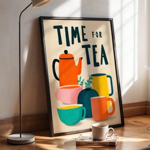 Op de afbeelding: Een ingelijste kunstprint met de tekst TIME FOR TEA in donkerblauwe letters, vergezeld van een kleurrijke illustratie van een oranje theepot, een roze kom, een groenblauwe kop, een donkerblauwe vaas, een gele mok en een oranje mok op een crèmekleurige achtergrond.