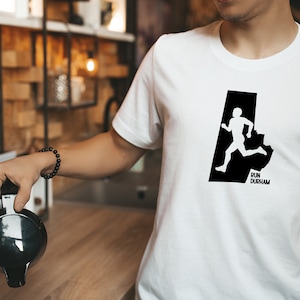 May include: White t-shirt with a black graphic of a runner inside a state outline, with the text "Run Durham" below. The shirt is being worn by a person.