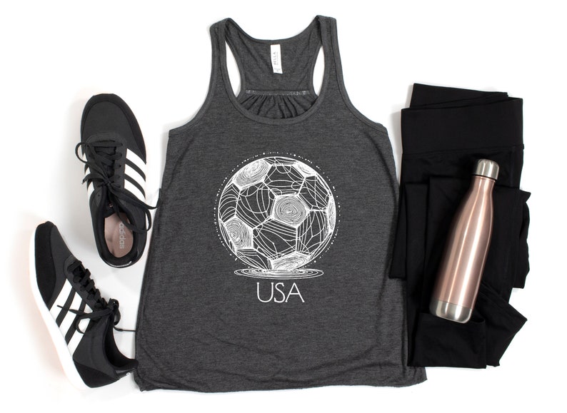 May include: A dark grey tank top with a white graphic of a soccer ball and the word "USA" underneath. The tank top is paired with black leggings and a rose gold water bottle.