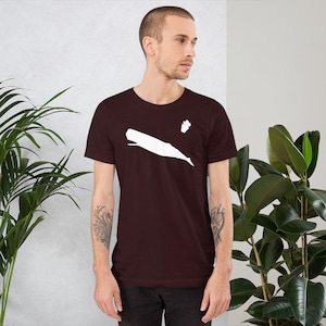 Puede incluir: Una camiseta marrón con una silueta blanca de una ballena y una pequeña flor en la parte delantera.