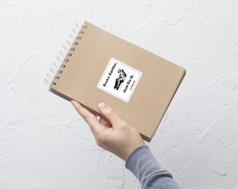May include: A brown notebook with a white sticker on the front. The sticker has a black and white illustration of a person in a car with the text "Brake Earlier. Just Do It." in black.