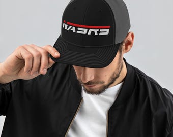 May include: A black and gray trucker hat featuring a black front panel with the white text "NASRD" underlined by a thin red horizontal line. The hat has a structured crown, a flat brim with stitched detailing, and a gray mesh back panel.