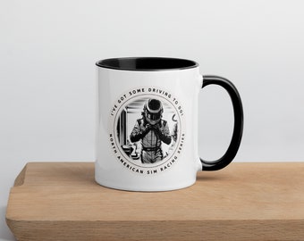May include: White ceramic mug with a black rim and handle. The mug features a black and white illustration of a race car driver with the text "I've got some driving to do. North American Sim Racing Series."