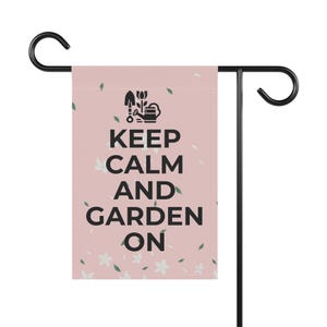 May include: A garden flag with a light pink background, featuring the text "KEEP CALM AND GARDEN ON" in black. The flag also has a black graphic of gardening tools and white flower and green leaf accents. The flag is attached to a black metal garden stake.