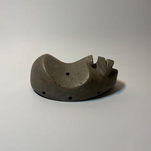 May include: A grey, abstract sculpture of a face, lying on its side. The sculpture has a rounded shape with carved features, including a nose and mouth. Several small holes are visible on the surface. The artwork is set against a white background.