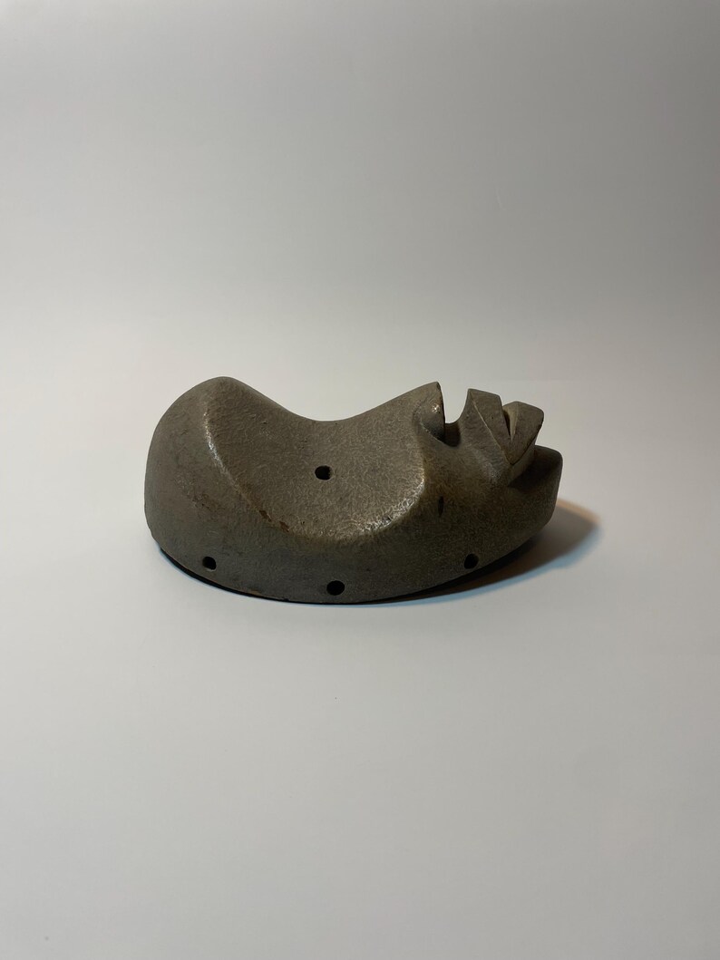 May include: A grey, abstract sculpture of a face, lying on its side. The sculpture has a rounded shape with carved features, including a nose and mouth. Several small holes are visible on the surface. The artwork is set against a white background.