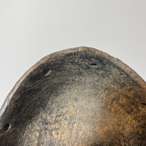 May include: Close-up of a weathered wooden mask with a curved shape. The mask has a dark, aged appearance with visible wear and tear, including small holes and cracks. The colour is a mix of dark brown and tan.