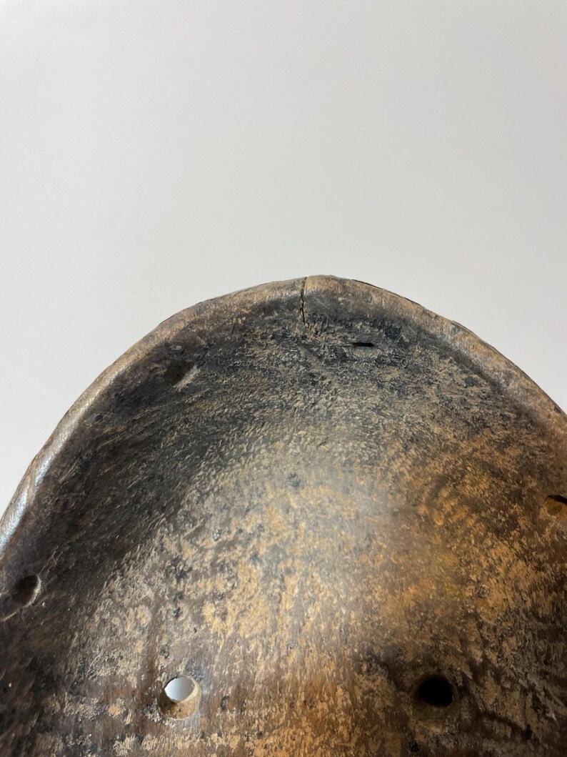 May include: Close-up of a weathered wooden mask with a curved shape. The mask has a dark, aged appearance with visible wear and tear, including small holes and cracks. The colour is a mix of dark brown and tan.