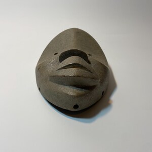 May include: A grey, carved mask with a stylised face. The mask has a pointed top and rounded bottom, with carved features including a nose, mouth, and eye holes. The mask is made of a textured material and is likely a decorative item.