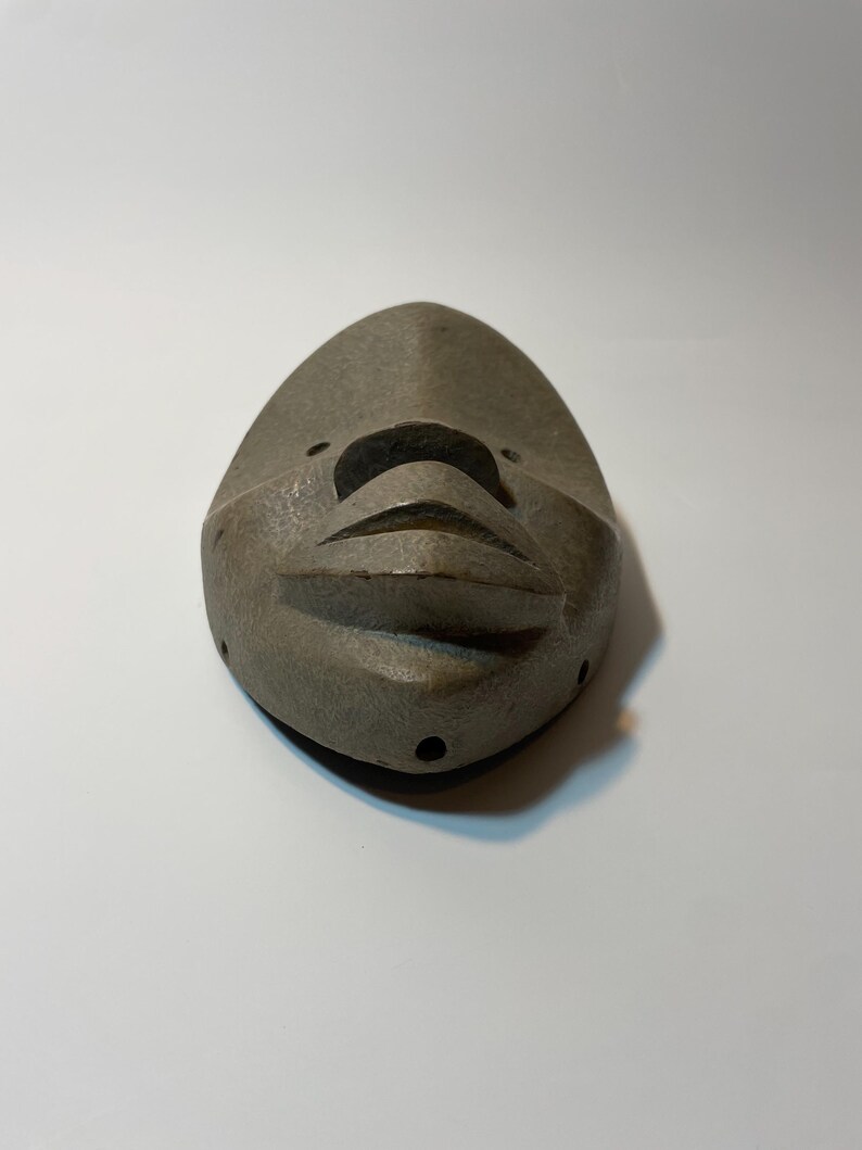 May include: A grey, carved mask with a stylised face. The mask has a pointed top and rounded bottom, with carved features including a nose, mouth, and eye holes. The mask is made of a textured material and is likely a decorative item.
