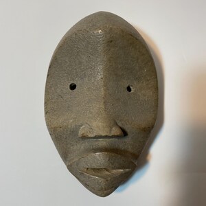 May include: A carved wooden mask in a muted grey colour with a symmetrical design. It features two eye holes, a small nose, and a defined mouth. The surface has a textured appearance.