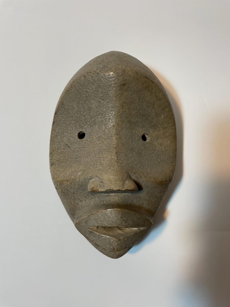 May include: A carved wooden mask in a muted grey colour with a symmetrical design. It features two eye holes, a small nose, and a defined mouth. The surface has a textured appearance.