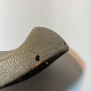 May include: Close-up of a weathered, grey object with a textured surface. The object has a curved shape with two small holes near the edge. The edges show signs of wear and tear, with some areas of discolouration.