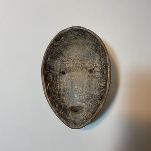 May include: Oval-shaped, weathered wooden mask with a face carved into it. The mask has two eye holes and a small, slit-like mouth. The colour is a mix of brown and grey, with visible wood grain.