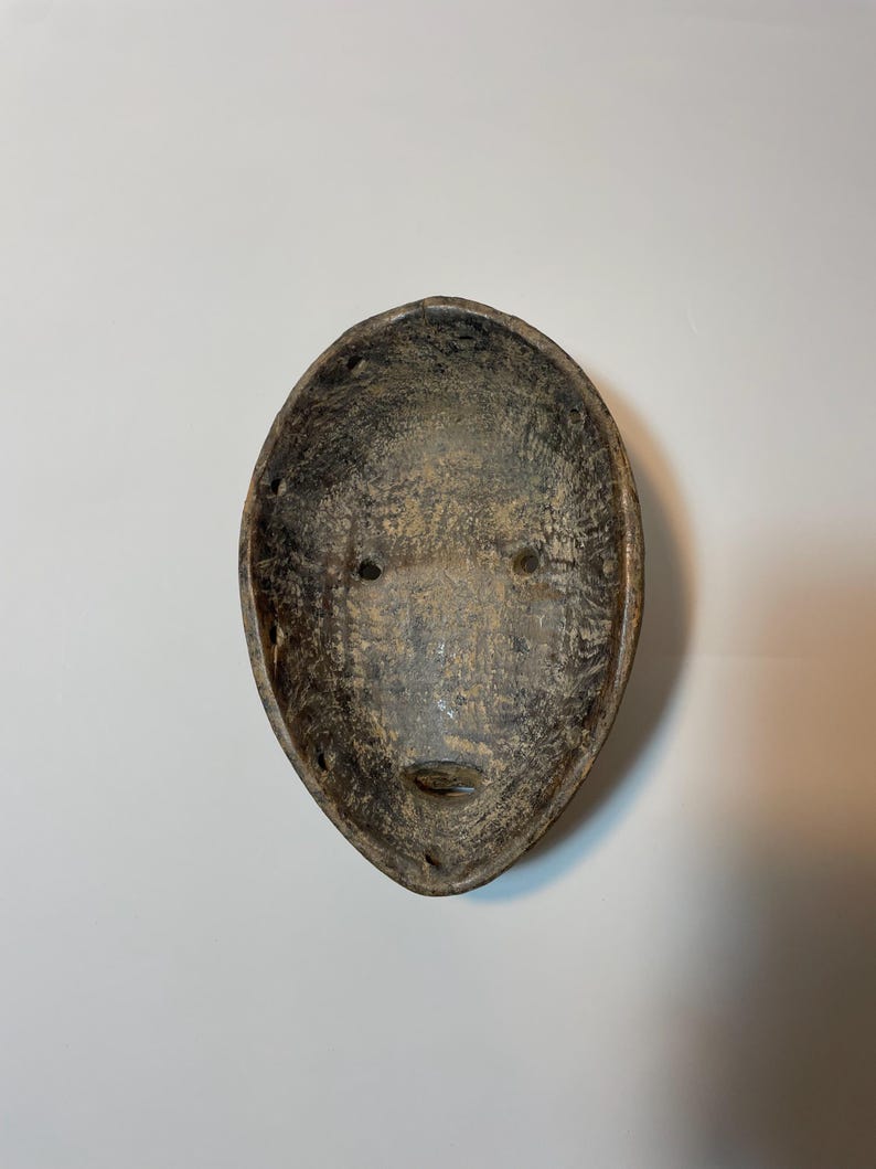 May include: Oval-shaped, weathered wooden mask with a face carved into it. The mask has two eye holes and a small, slit-like mouth. The colour is a mix of brown and grey, with visible wood grain.