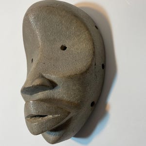 May include: A grey, carved mask depicting a human face. The mask features a pronounced nose, full lips, and a rounded forehead. Several small holes are drilled around the eye and ear areas. The mask is likely made of stone or a similar material.