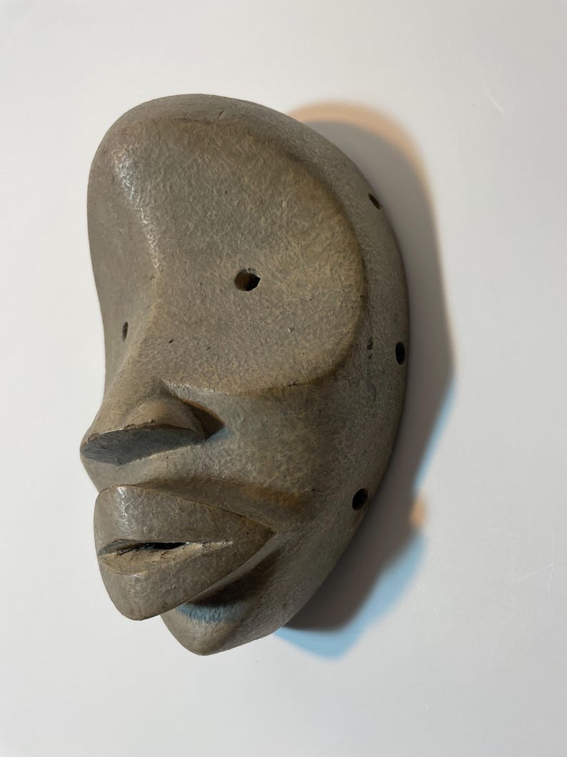 May include: A grey, carved mask depicting a human face. The mask features a pronounced nose, full lips, and a rounded forehead. Several small holes are drilled around the eye and ear areas. The mask is likely made of stone or a similar material.