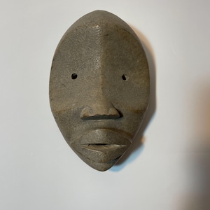 May include: A carved, grey wooden mask with a stylized face. The mask has a smooth, oval shape with two small eye holes, a prominent nose, and a closed mouth. The surface has a textured appearance.